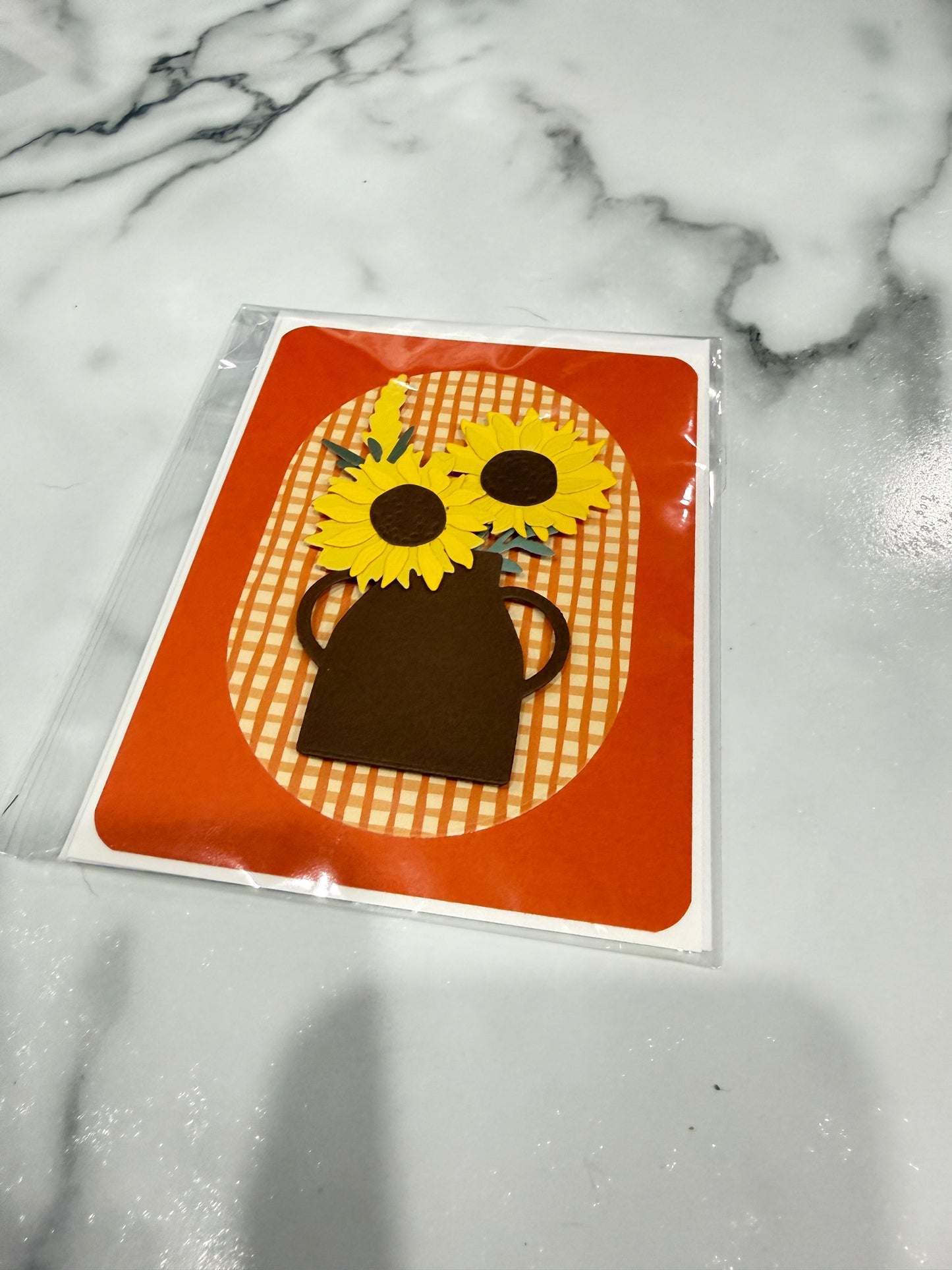 A square greeting card featuring a sunflower and vase design, sealed in a clear plastic sleeve and placed on a white marble surface.