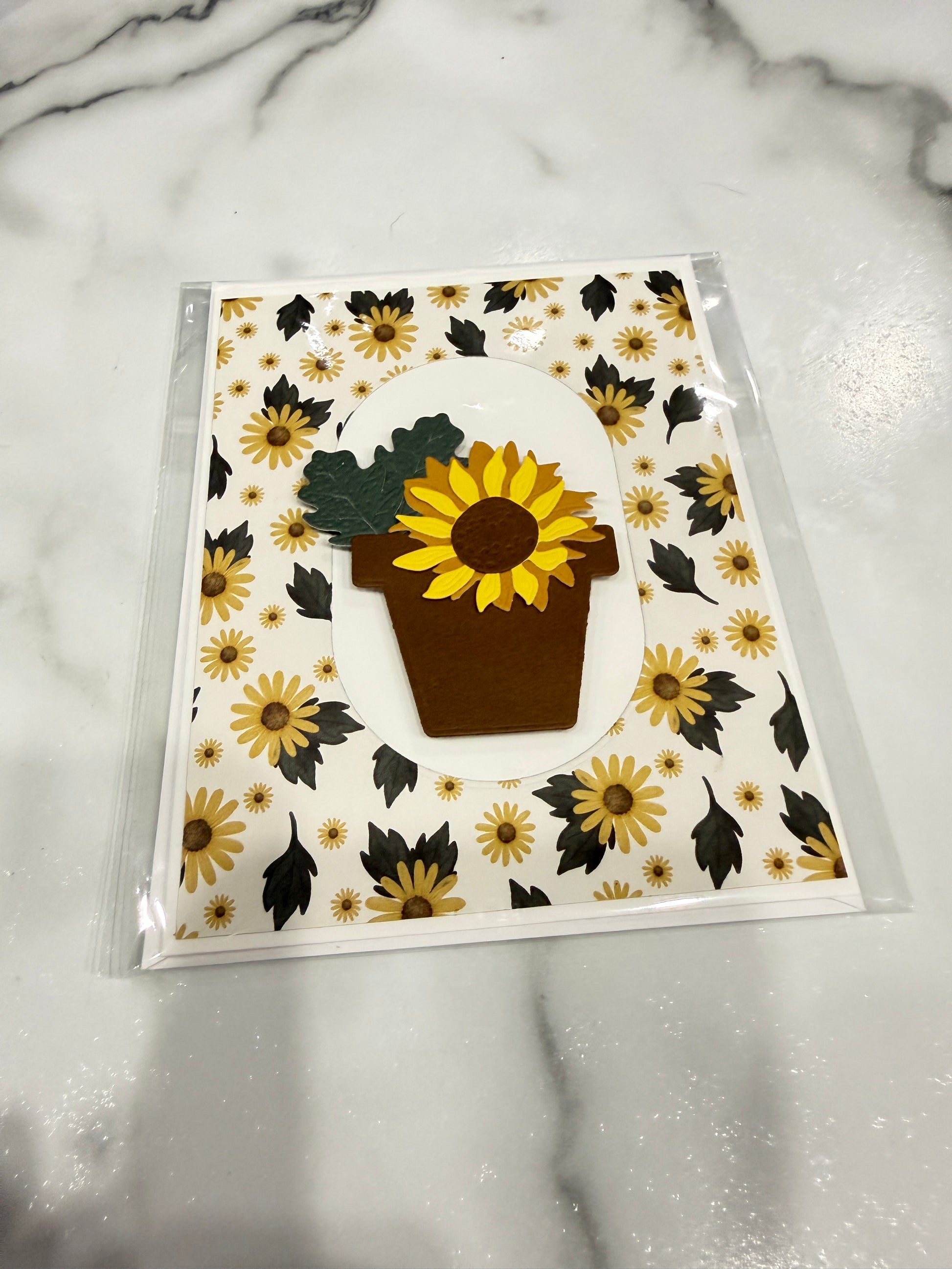 A handmade paper card featuring a central brown pot with a yellow sunflower and green leaves, surrounded by a white background patterned with yellow sunflowers and black leaves. The card is sealed in a clear plastic sleeve and placed on a white marble surface.