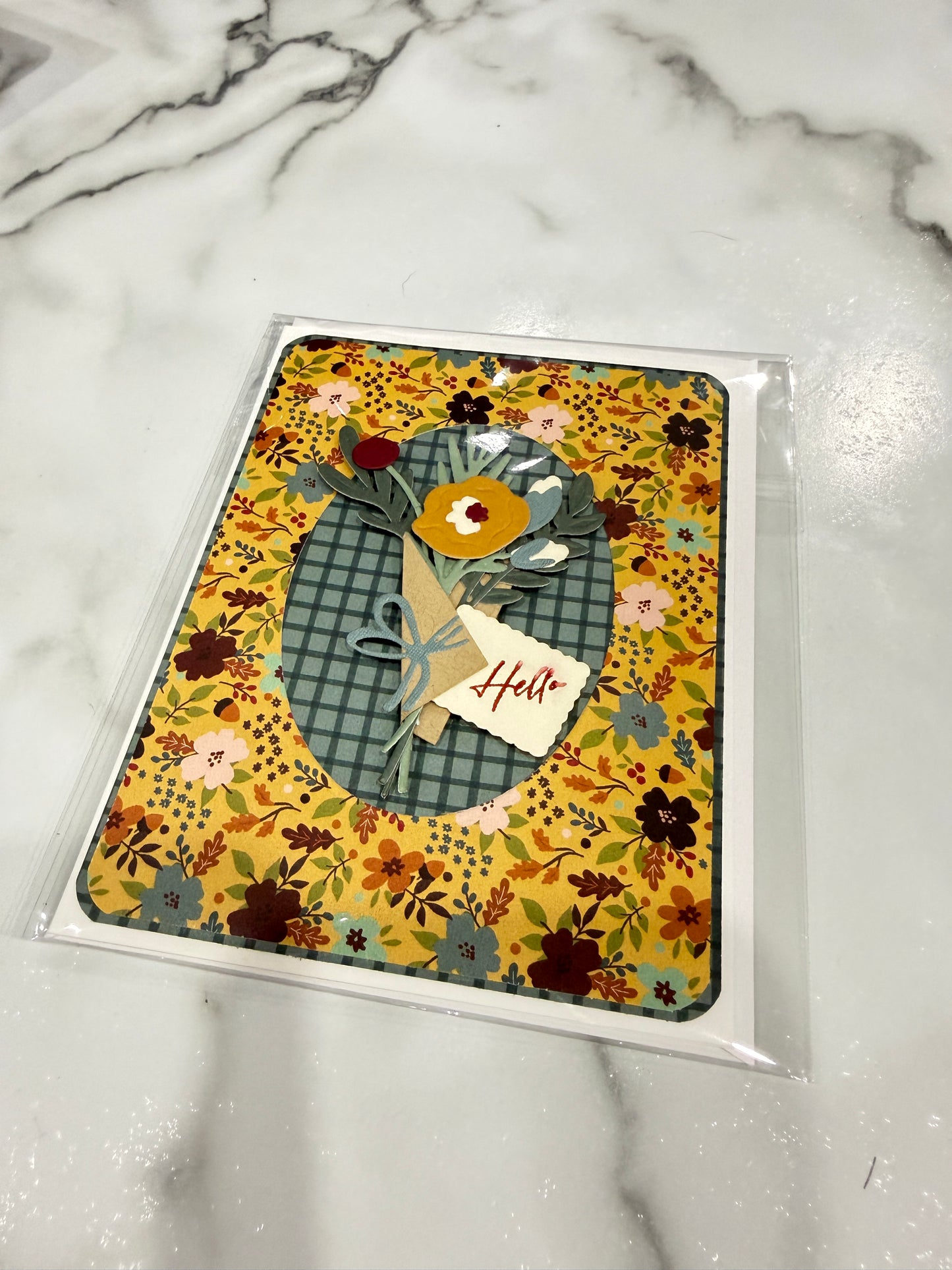 A handmade greeting card in clear plastic packaging, featuring a yellow floral background, a blue-green plaid oval, and a small paper flower with a 'Hello' tag, resting on a white marble surface.