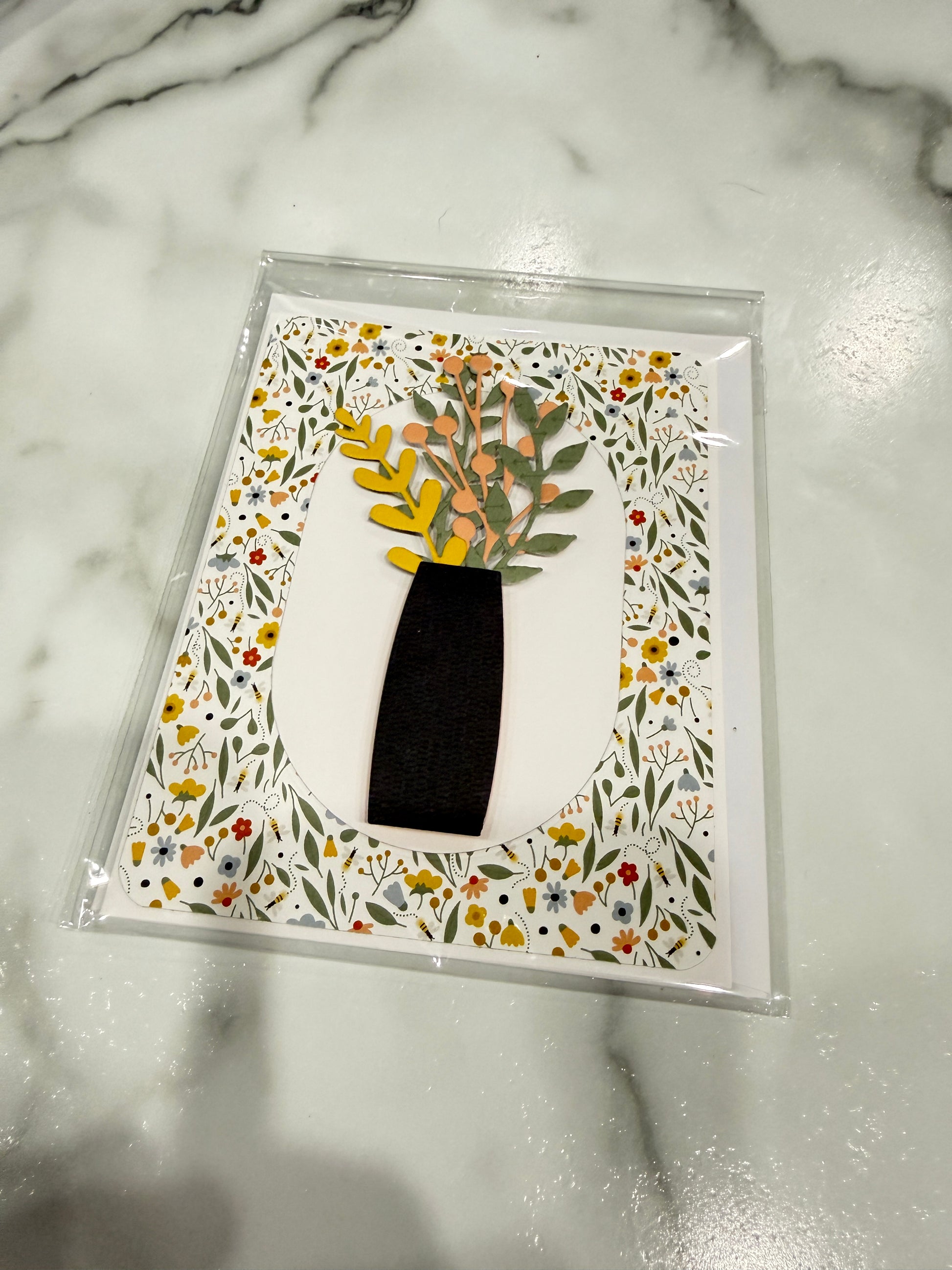 A square decorative art print or card in a clear plastic sleeve, featuring a black vase with yellow, orange, and green floral stems on a white background, surrounded by a dense multicolor floral border. The print is photographed on a white marble surface.