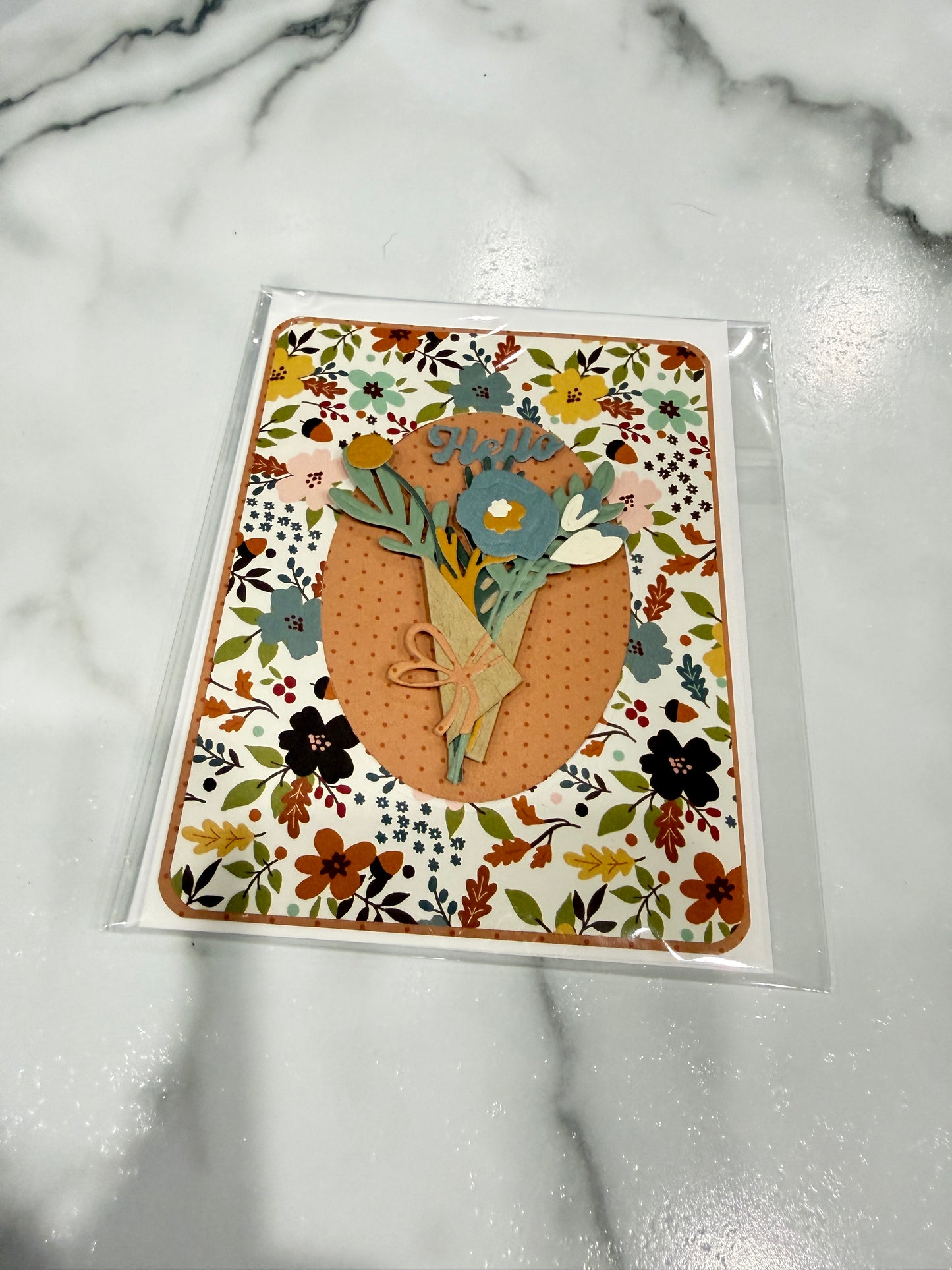 A handmade floral greeting card in clear plastic packaging, featuring a central orange oval with layered paper flowers and leaves on a white marble background.