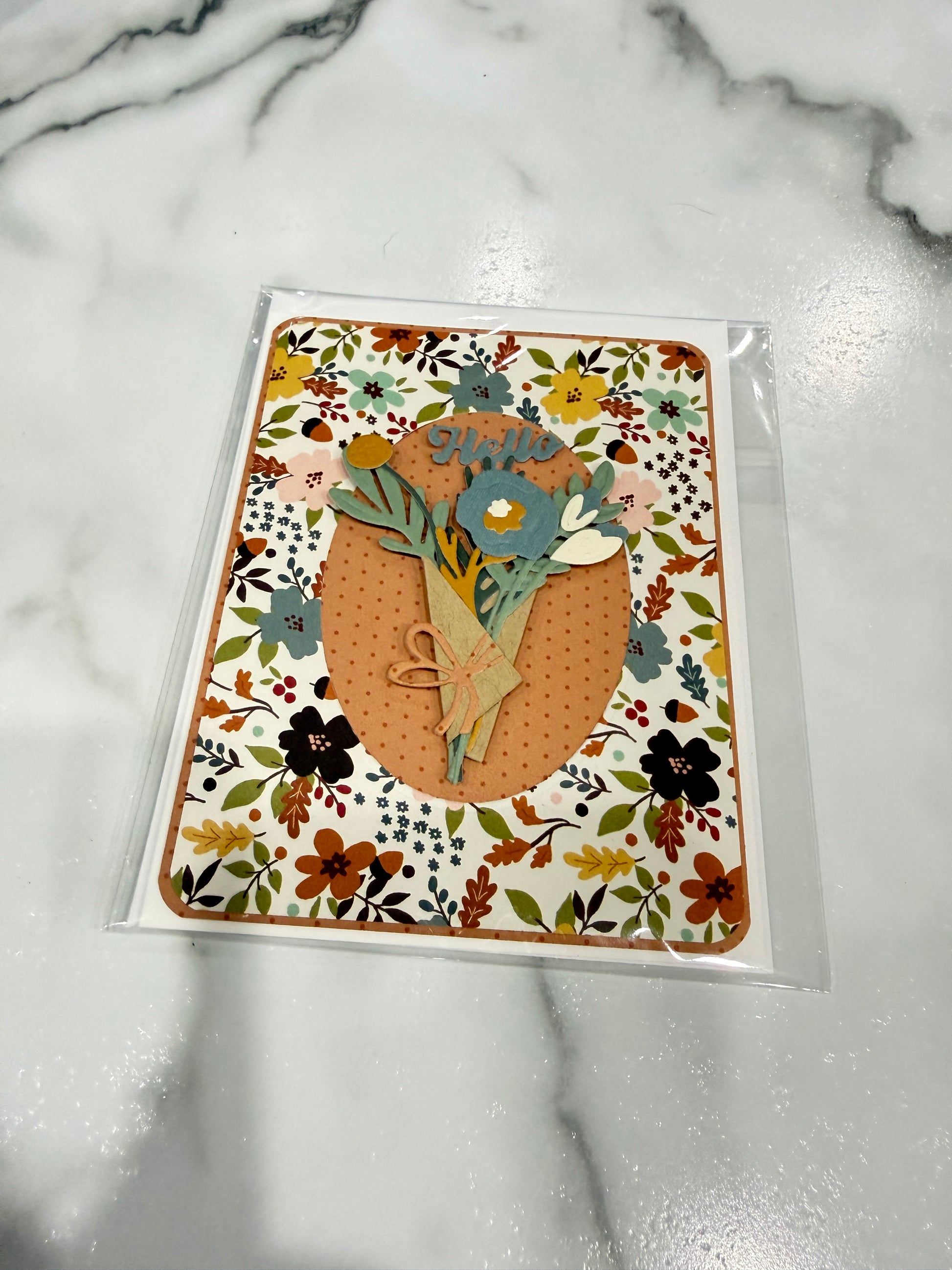A handmade floral greeting card in clear plastic packaging, featuring a central orange oval with layered paper flowers and leaves on a white marble background.
