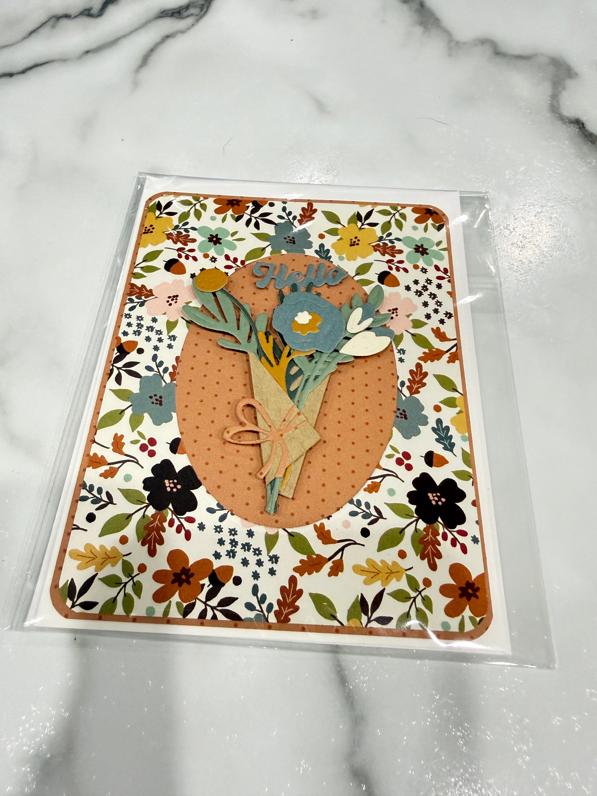 A handmade greeting card in clear plastic packaging, featuring a floral patterned border and a central layered design with a peach oval, green leaves, and a blue flower. The card is placed on a white marble surface.