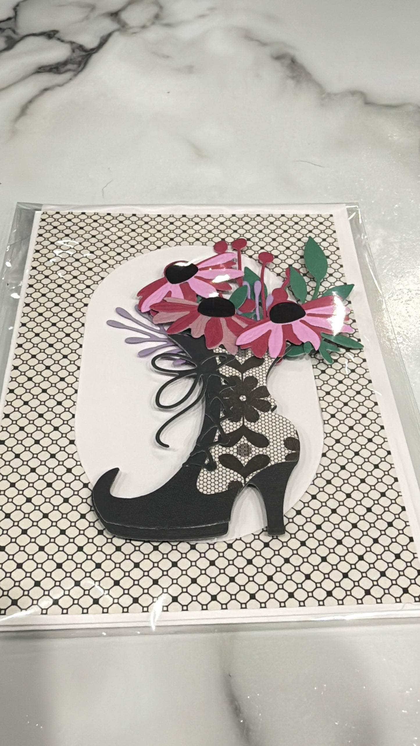 A decorative art print or card featuring a black high-heeled shoe filled with pink flowers and green leaves, placed on a patterned black-and-white background, photographed on a marble surface.