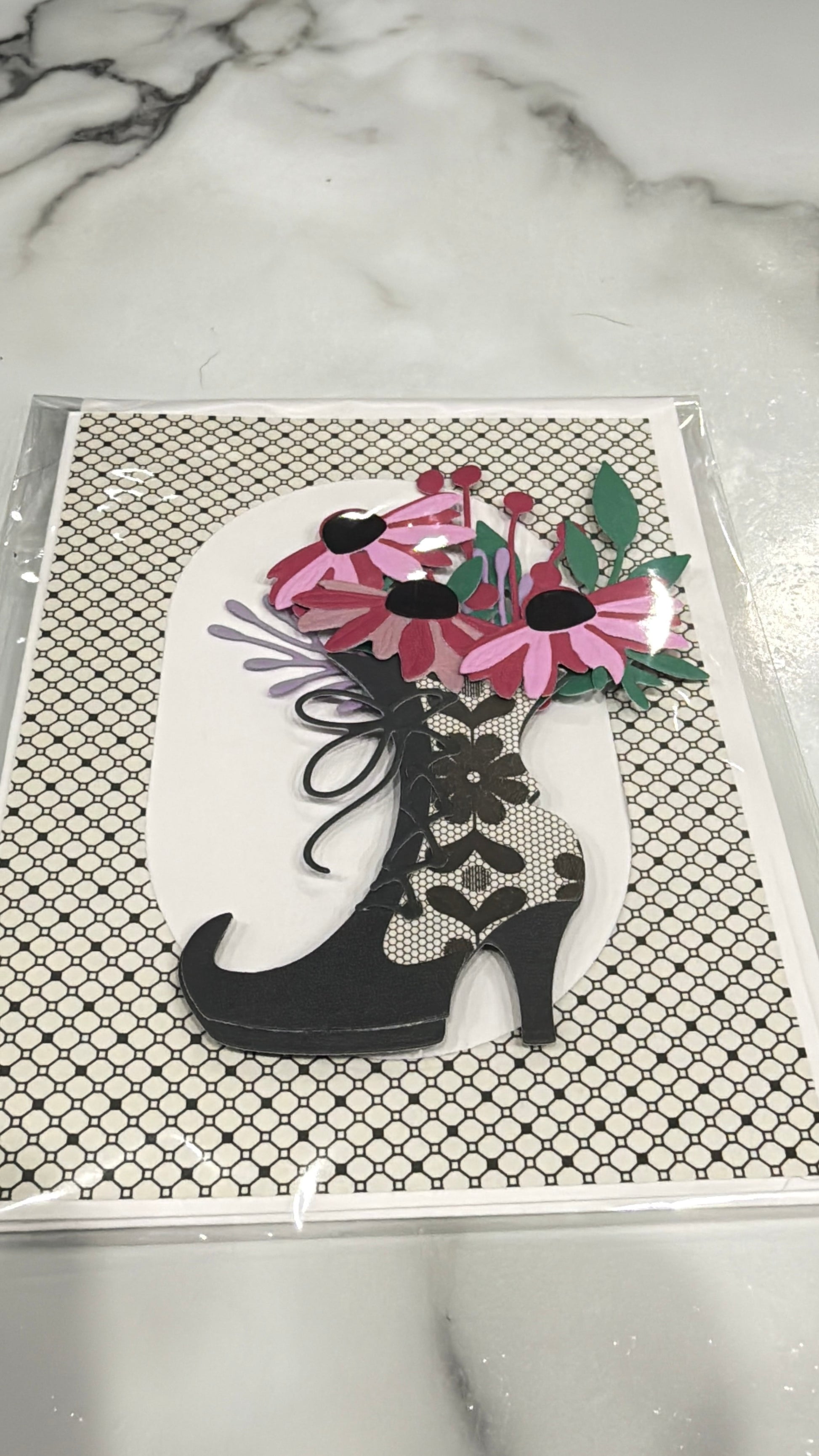 A decorative art print or card featuring a black high-heeled shoe filled with pink flowers and green leaves, placed on a patterned black-and-white background, photographed on a marble surface.
