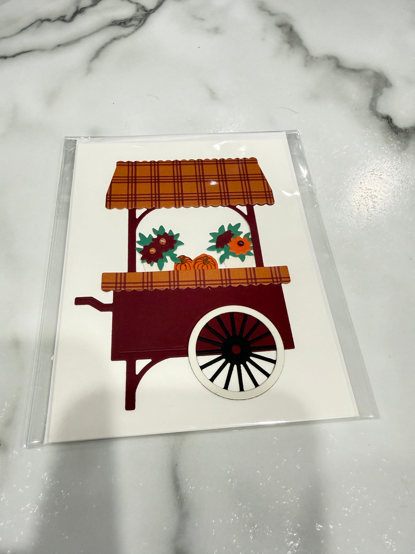 A square white card featuring a stylized autumn market cart illustration in orange, red, green, and black, sealed in a clear plastic sleeve and placed on a white marble surface.