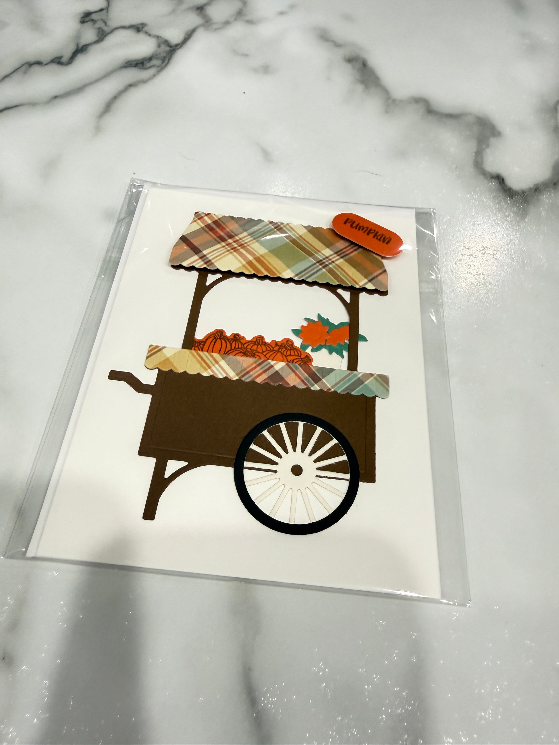 A square white card featuring a stylized autumn market cart illustration with plaid awning, orange pumpkins, and a small orange tag reading 'Fruitful', photographed on a white marble surface inside a clear sleeve.