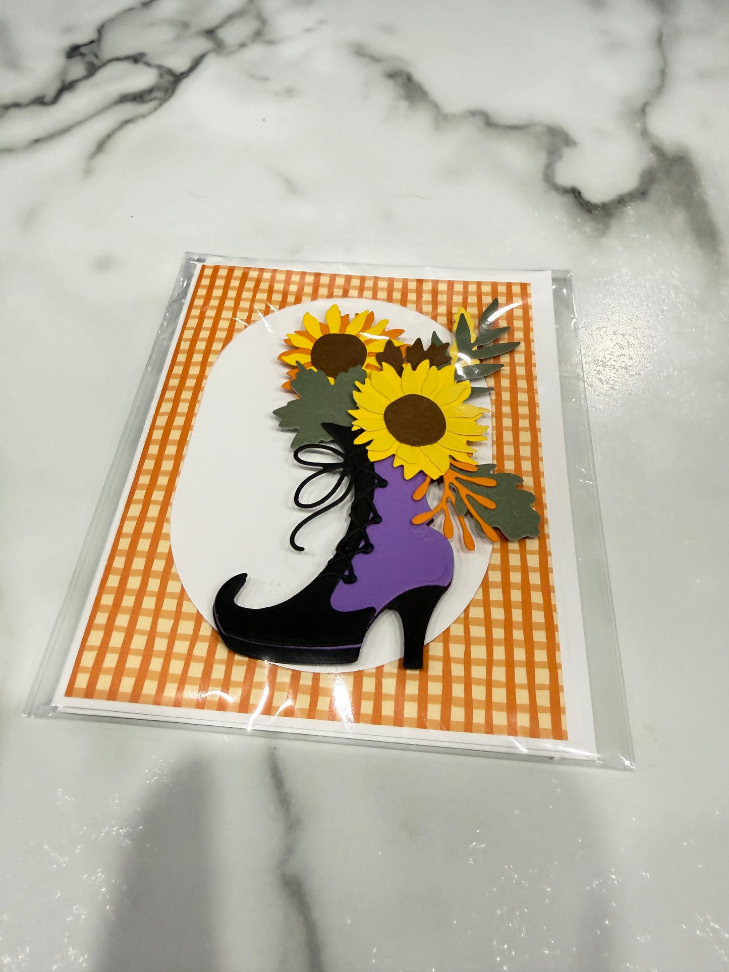 A handmade layered paper craft card featuring a black high-heeled boot filled with purple fabric, decorated with yellow sunflowers and green leaves on an orange and white gingham background. The card is sealed in a clear plastic sleeve and placed on a white marble surface.