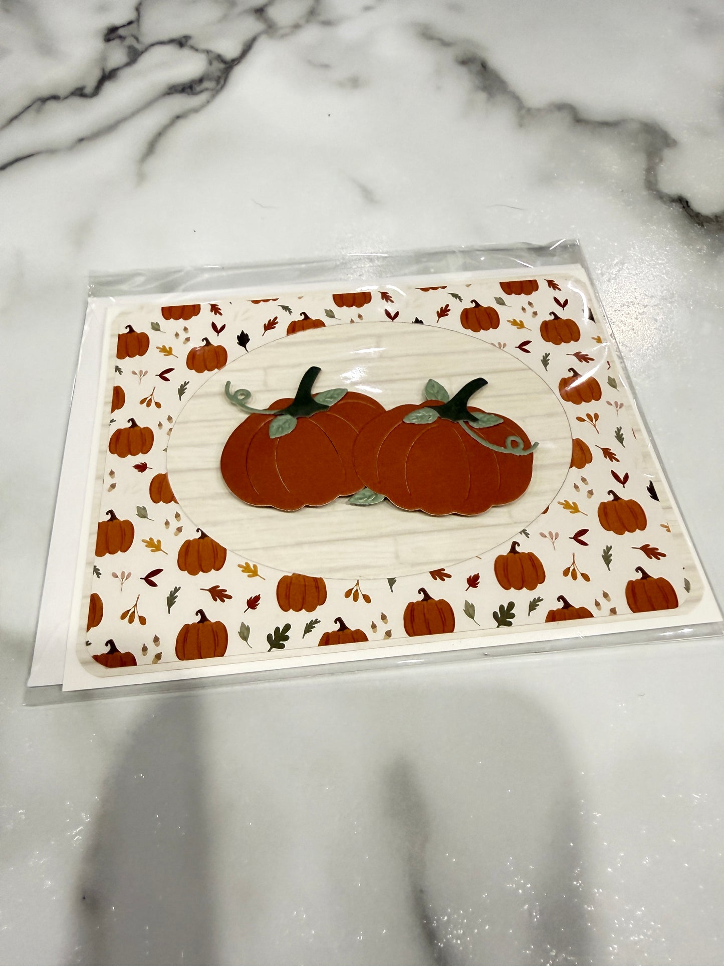 A square white card with a fall pumpkin pattern, featuring two layered orange pumpkins in the center, photographed on a white marble surface inside a clear plastic sleeve.
