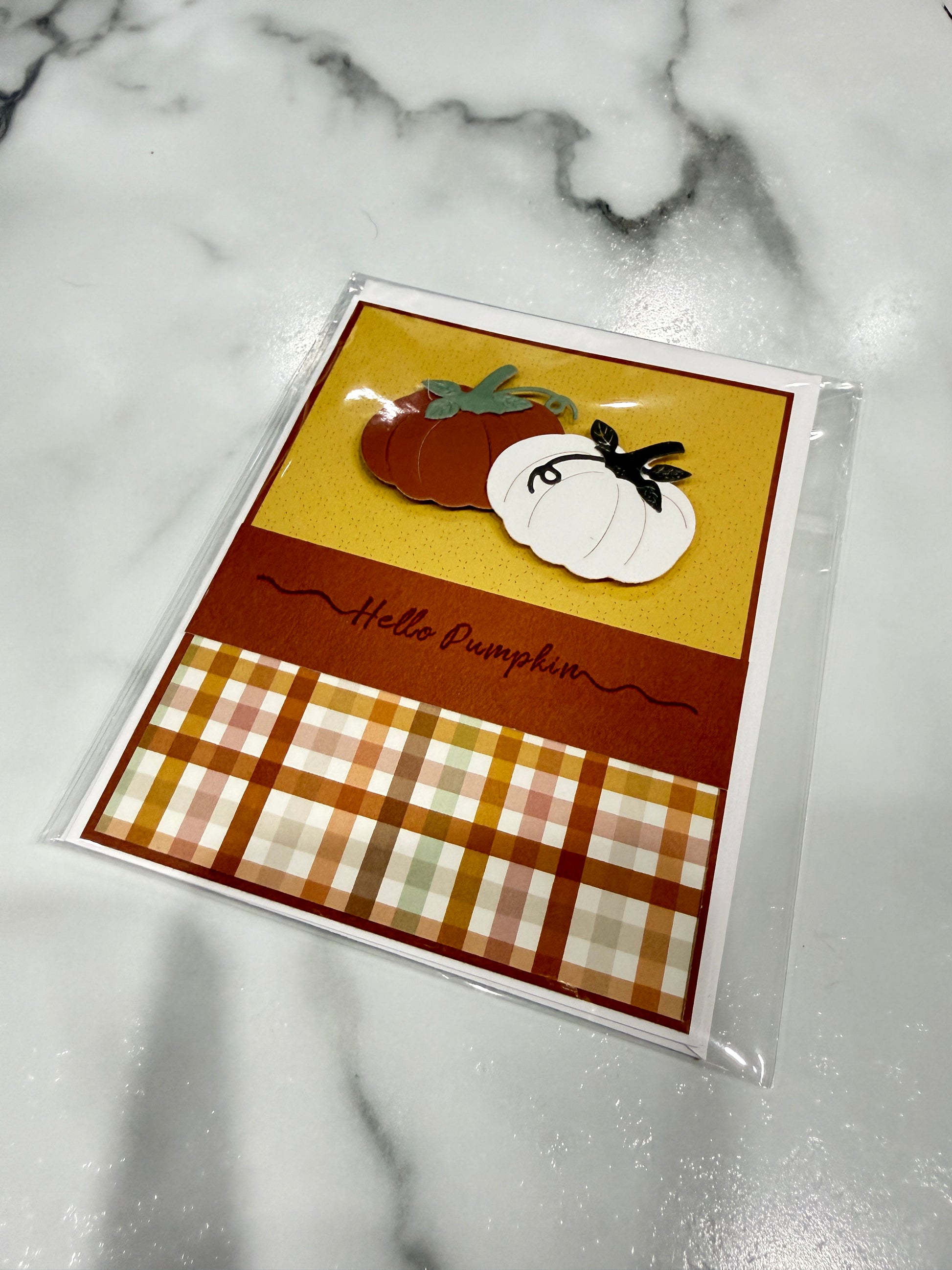 A handmade autumn greeting card in clear plastic packaging, featuring layered paper pumpkins, a gold background, a brown band with 'Hello Pumpkin' in script, and an orange, beige, and white plaid pattern at the bottom, displayed on a white marble surface.