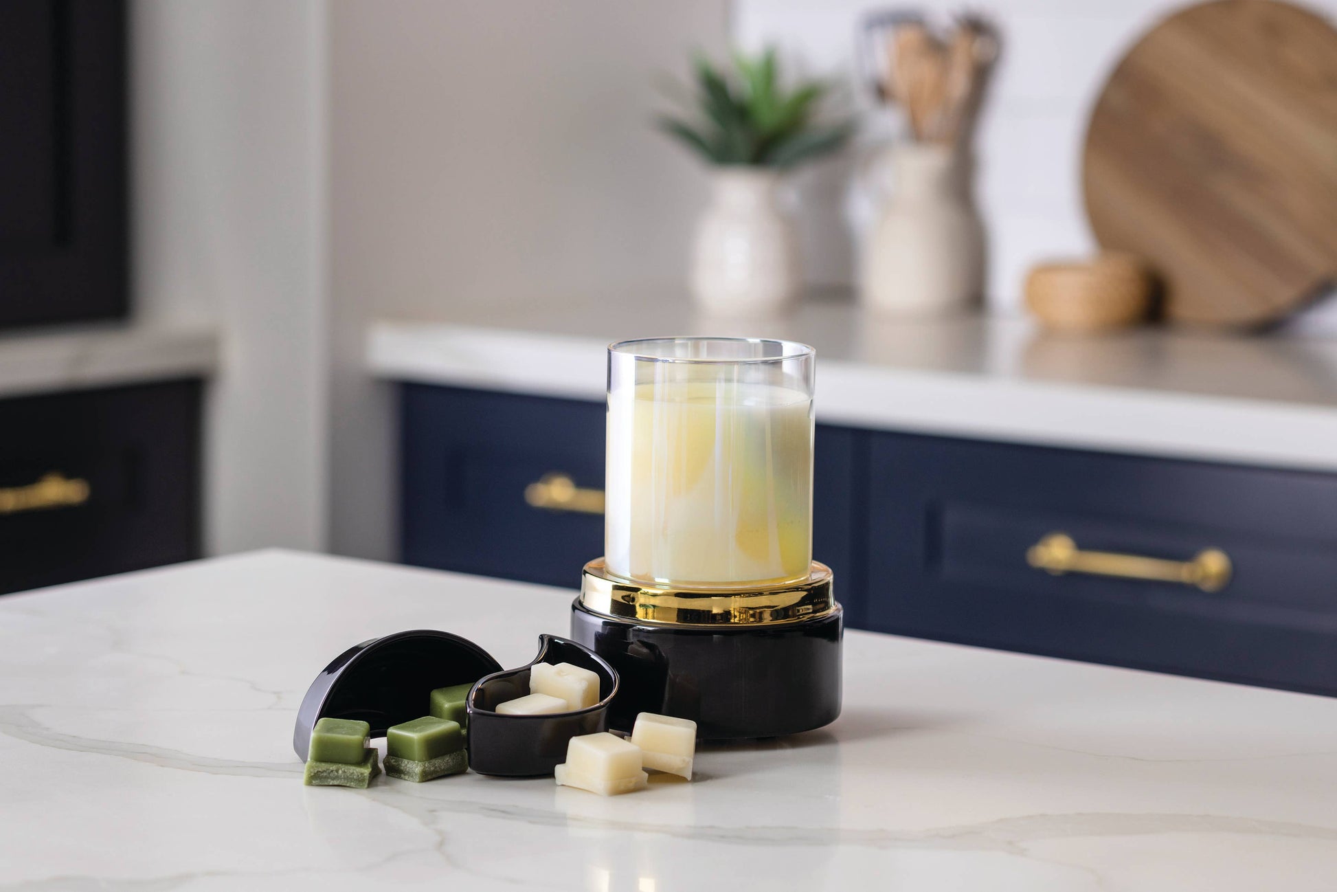A black and gold wax melt warmer with a clear glass jar containing a yellow and white marbled wax melt, shown on a white marble kitchen counter with wax melts and green wax cubes nearby.