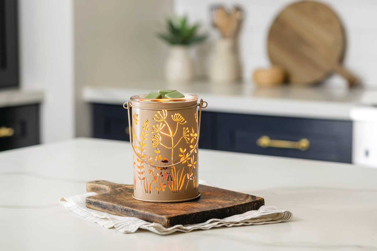 A copper-colored metal tea light holder with floral cutout patterns sits on a dark wooden cutting board on a white marble kitchen countertop, with a blurred kitchen background.