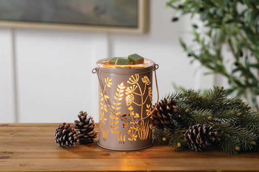 A metal candle warmer lantern with a warm gold botanical pattern, lit from within, sits on a wooden table surrounded by pinecones and evergreen branches.