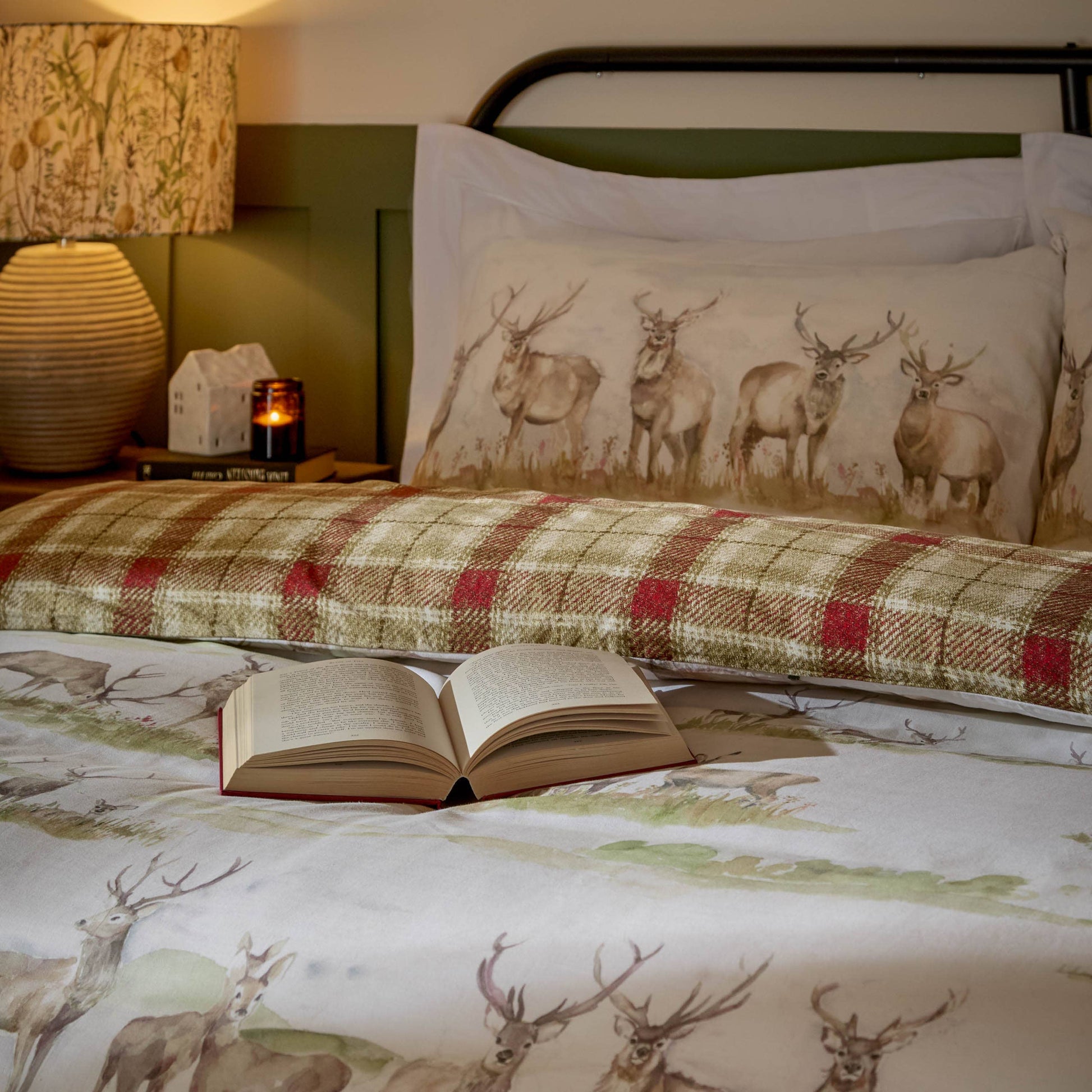 Cozy woodland-themed bedding featuring illustrated deer, a coordinating plaid bolster pillow, and soft neutral tones styled on a bed with an open book.