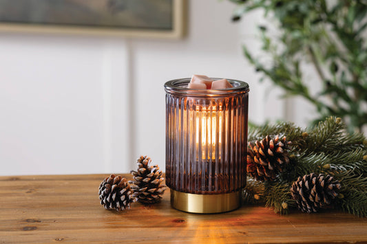 A ribbed amber glass candle holder with a gold-toned base, lit with a warm candlelight glow, styled on a wooden table with pinecones and evergreen branches.