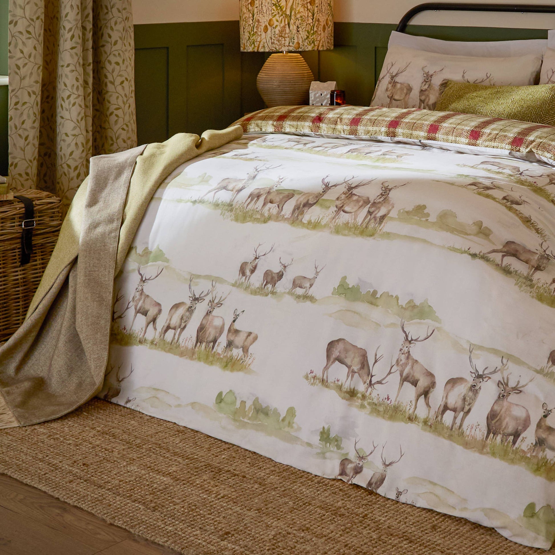 A made bed in a styled bedroom featuring a duvet cover with a watercolor-style deer herd pattern in muted green, beige, and brown tones on an off-white background. Matching patterned pillows are visible, along with a tan throw blanket draped over the foot of the bed and a wicker basket beside it.