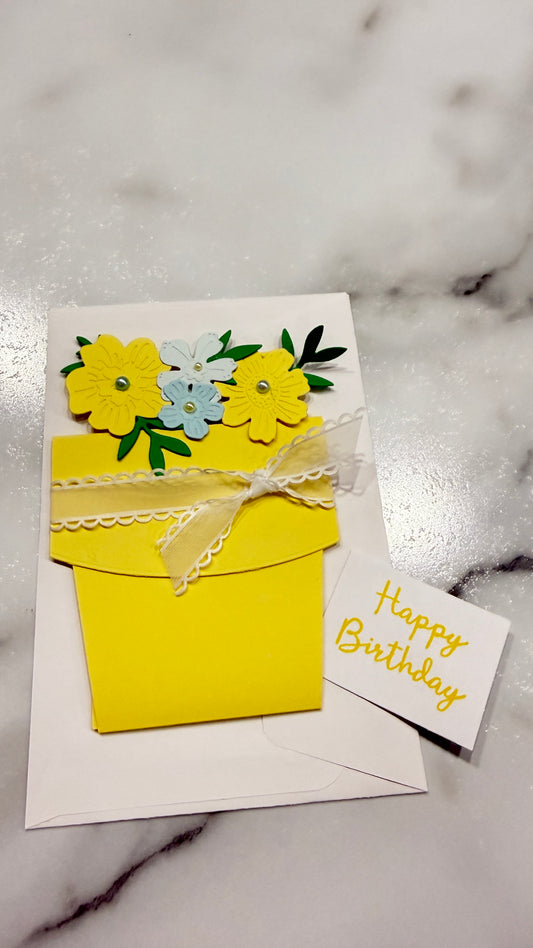 A handmade yellow envelope-style birthday card with layered paper flowers in yellow, light blue, and green, tied with a white lace ribbon, shown on a white marble surface next to a small card reading 'Happy Birthday'.