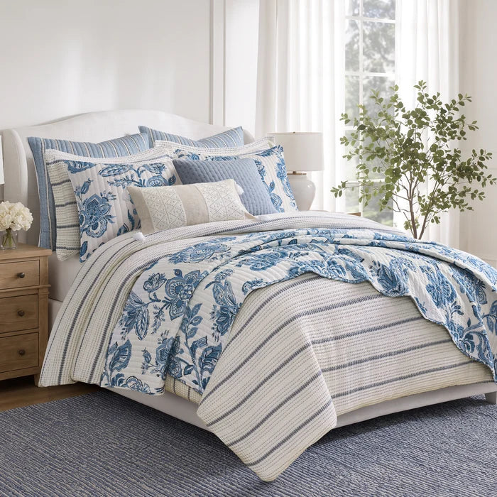 Blue and ivory waffle stripe comforter layered with blue floral quilt and coordinating pillows in a bright coastal style bedroom.
