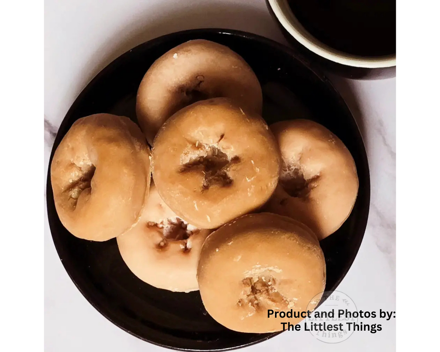 Six glazed donuts arranged on a black plate, photographed from above on a light marble-like surface. A partial white cup with dark liquid is visible at the top right, and a watermark at the bottom right reads 'Product and Photos by: The Littlest Things'.
