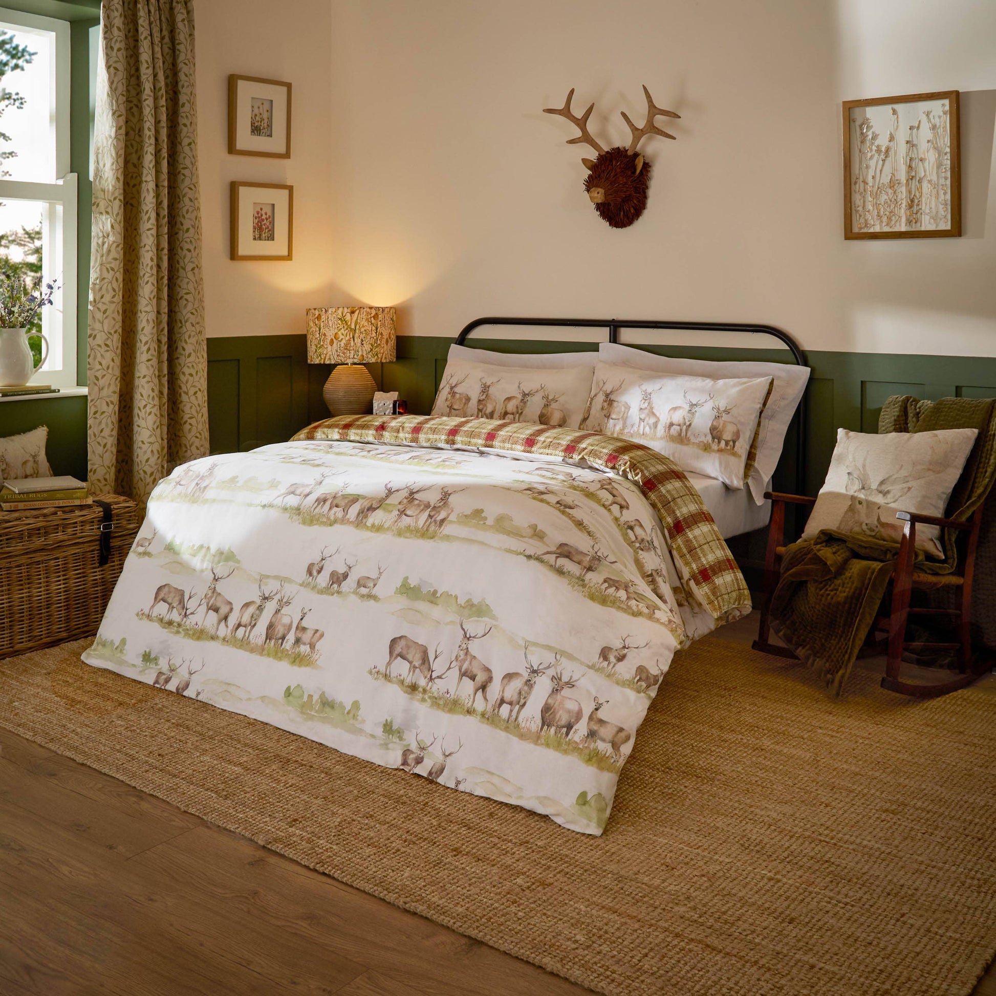 A rustic bedroom scene featuring a bed dressed in a white duvet cover and pillowcases printed with a deer and elk safari pattern, styled with a green and brown wood-paneled wall, a lit bedside lamp, and a wooden chair.
