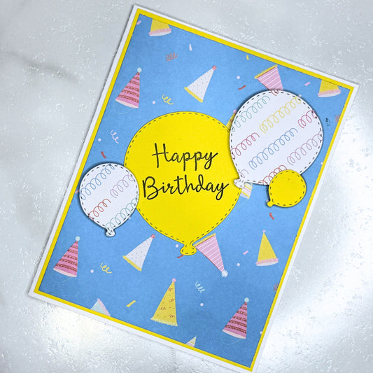 A handmade square birthday card featuring a blue background with a yellow border, decorated with illustrated party hats, balloons, and the words 'Happy Birthday' in black script inside a large yellow balloon.