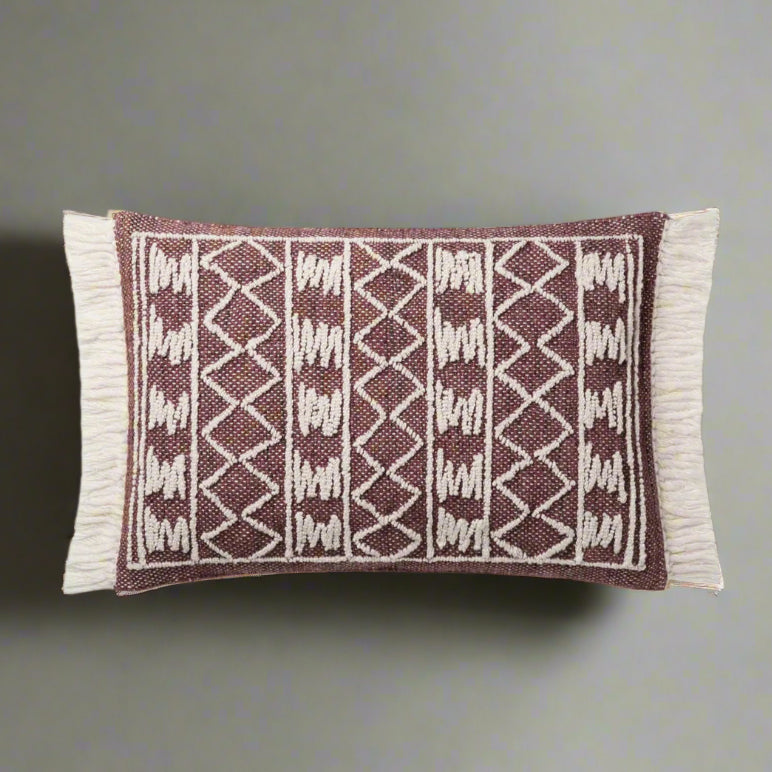 A rectangular lumbar pillow with a warm burgundy background and an ivory geometric pattern, featuring textured weave and fringe detailing along the edges.