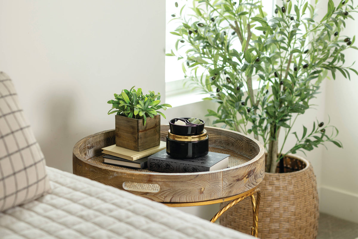 A round wooden side table with a gold metal frame, styled in a bright room next to a bed with a plaid pillow and a large potted olive tree.