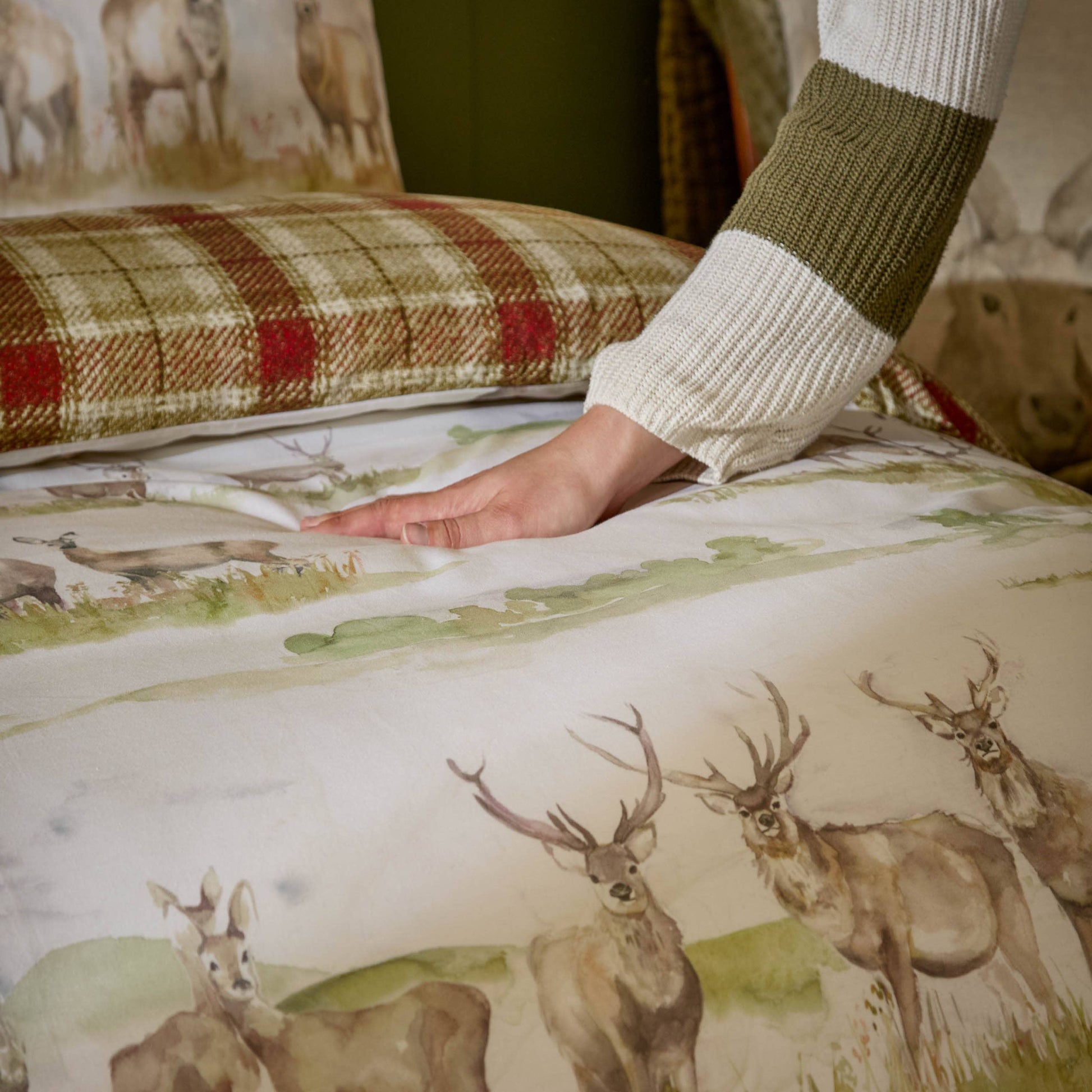 A close-up of a person's hand resting on a deer-print bedding sheet, with a striped knit sleeve visible. The sheet features a scenic woodland landscape with several deer, and a plaid pillowcase is visible in the background.